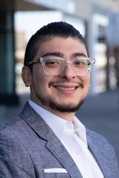 Dr. Paul Martinez smiling, wearing a gray suit, standing outdoors in an urban setting.